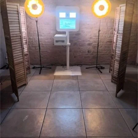 Borne photobooth blanche sur pied, encadrée de deux projecteurs et de paravents en bois